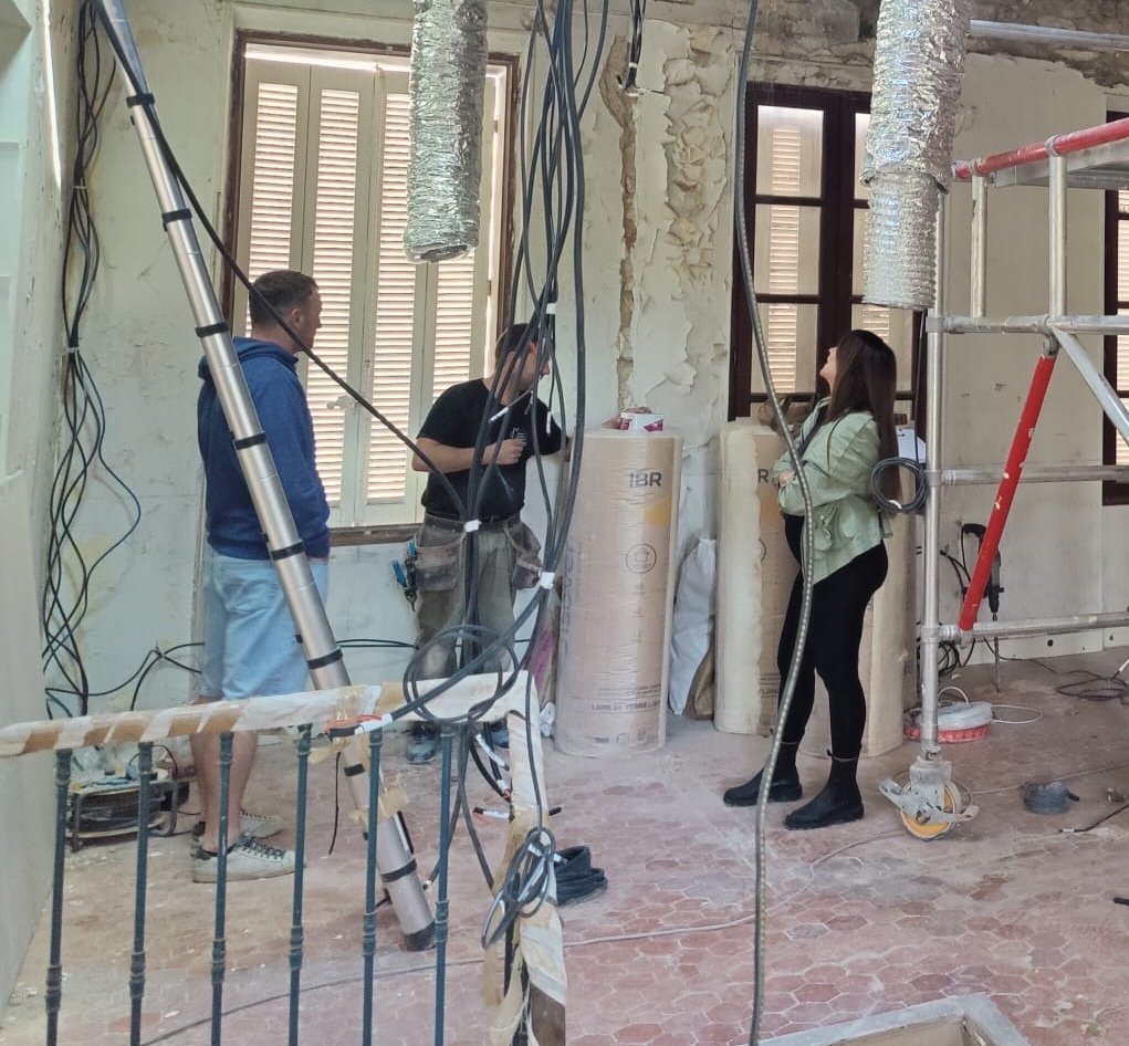 Une architecte et des ouvriers discutent dans une pièce en rénovation, entourées de matériaux de construction et de câbles électriques.