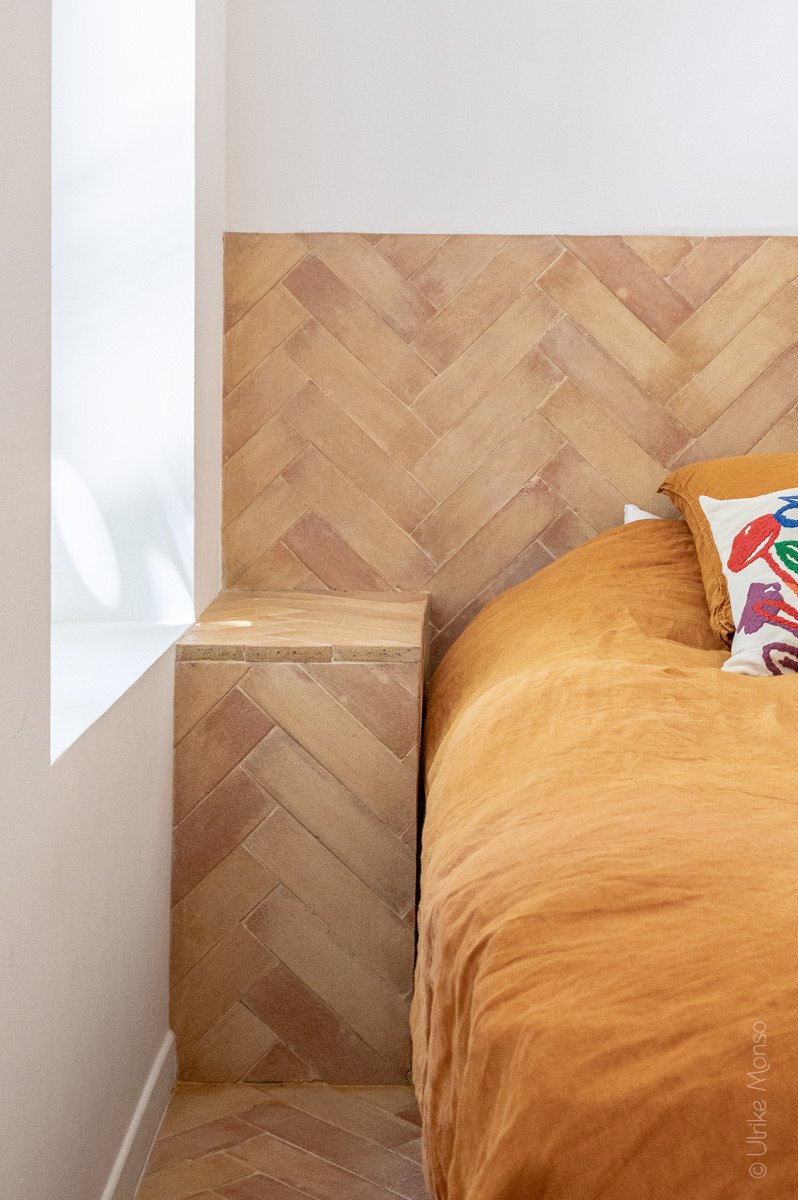 Coin de chambre avec un mur en briques en chevron, un lit aux draps orange et un coussin coloré.