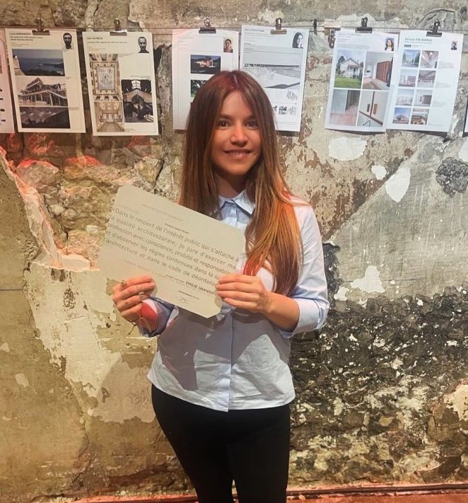 Une jeune femme tient son diplome d'architecte dans les mains devant des affiches dans un espace artistique.