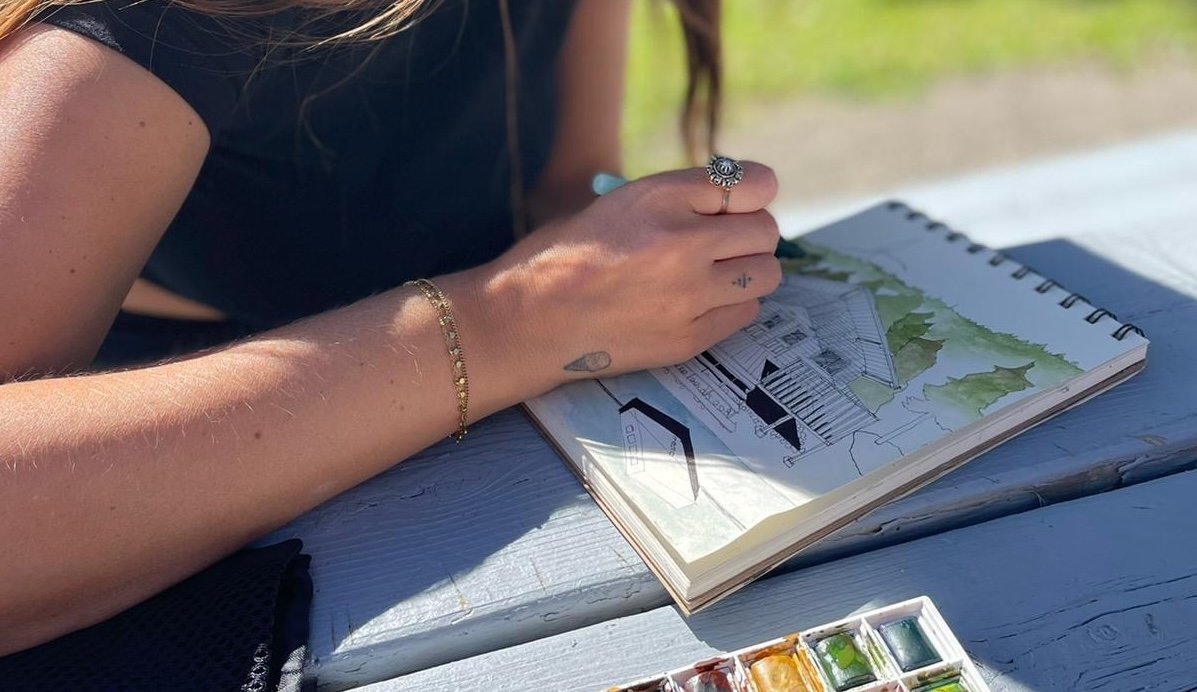 Une main dessine une maison dans un carnet de croquis, avec une palette de couleurs à côté sur une table en bois.