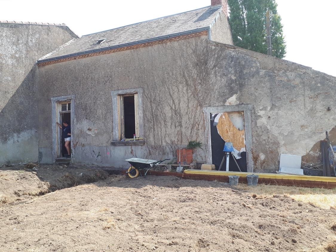 Maison en rénovation avec des fenêtres endommagées, une porte ouverte et des outils de construction à l'extérieur.