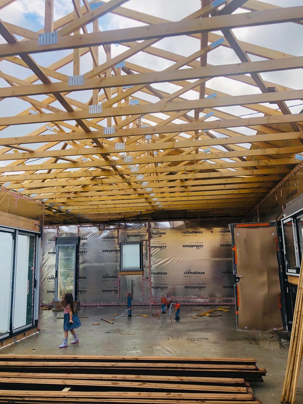Intérieur d'une construction avec une charpente en bois, une enfant explore l'espace, et des matériaux de construction sont visibles.